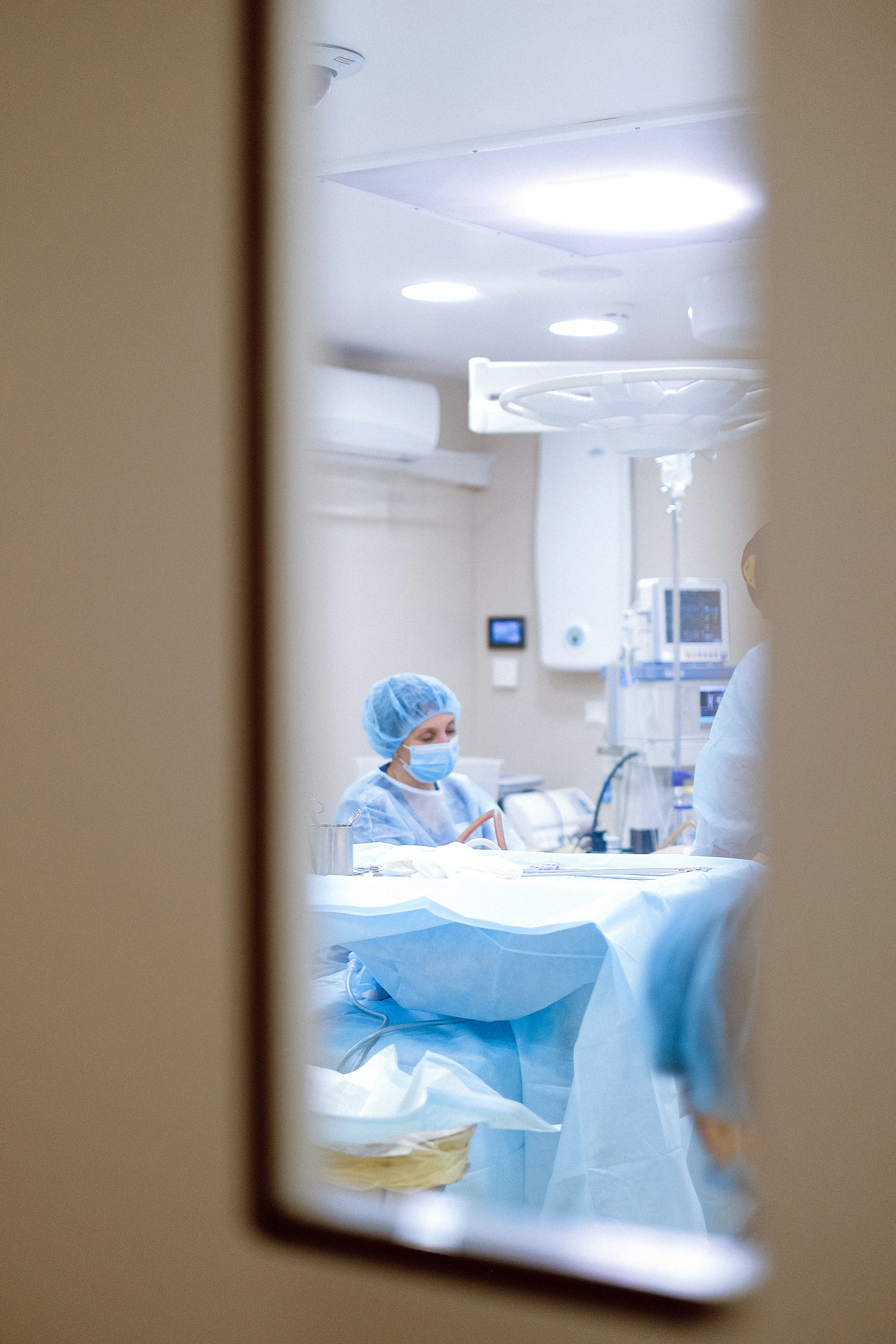 View through a narrow door window into an operating theatre, where a nurse in blue surgical scrubs, mask, and cap sits beside a prepared operating table under clinical lighting, surrounded by medical equipment in a clean, sterile environment.