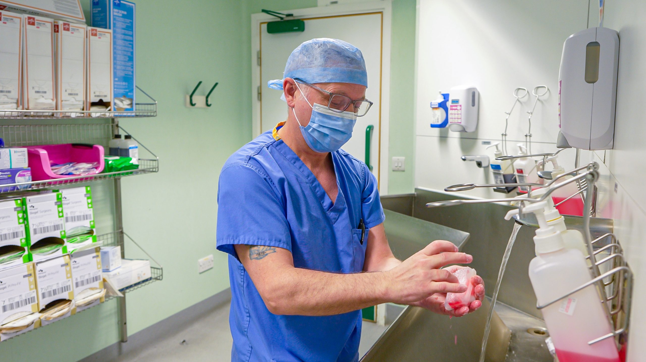 Assistant Theatre Practitioner in blue scrubs, mask, and cap washing hands at a clinical sink in a hospital scrub room, with medical supplies and equipment visible in the background.