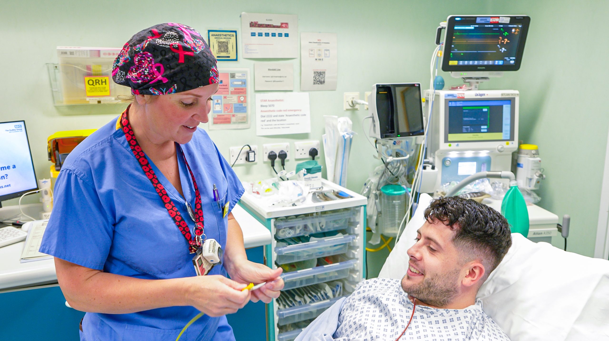 A Theatre Support Worker in blue scrubs and a patterned surgical cap smiles while holding a piece of medical tubing, speaking to a male patient lying in a hospital bed. The patient, wearing a hospital gown, looks up at her with a relaxed expression. Medical equipment, monitors, and supplies are visible around the bedside in a clinical hospital setting.