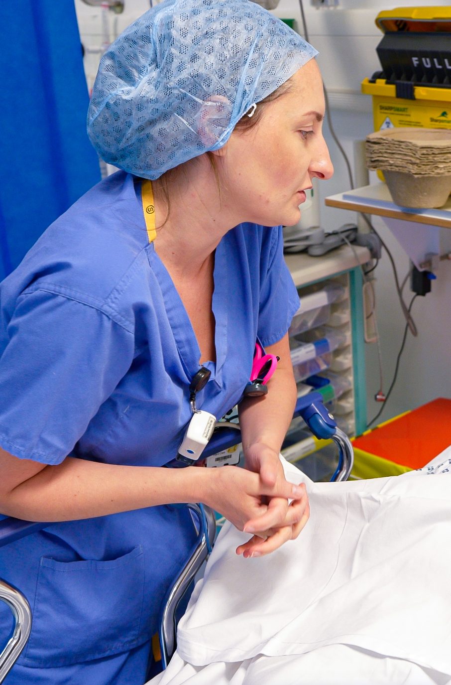 Nursing Associate in blue surgical scrubs and a hair cover sits beside a patient bed in a clinical setting, leaning forward with hands clasped, appearing focused and attentive. Medical equipment and storage trays are visible in the background.