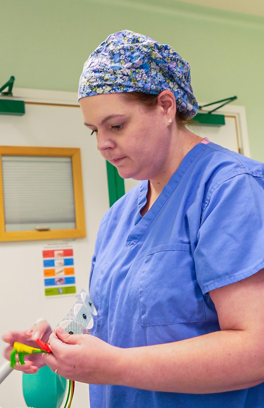 An Operating Department Practitioner in blue scrubs and a patterned surgical cap prepares equipment in a clinical setting, standing in front of a hospital door with a small window and signage visible.