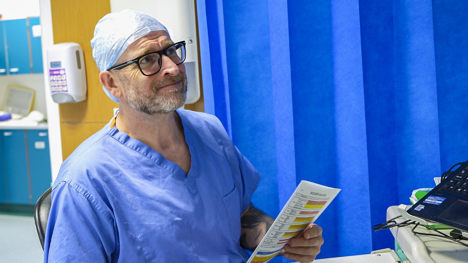 Male recovery practitioner in an operating theatre wearing blue scrubs, a surgical cap, and glasses, holding and reviewing a printed checklist or notes. Medical equipment and monitors are visible beside him, with a blue privacy curtain in the background.