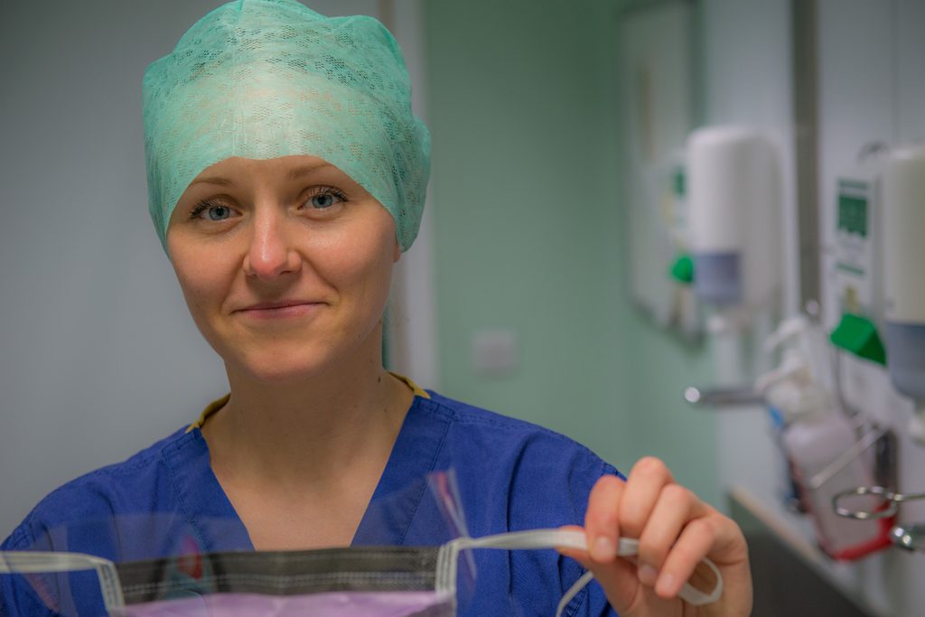 Theatre nurse in an operating room holding a sterile surgical mask in gloved hands, with medical equipment and clinical surroundings visible in the background.